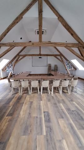 Salle mansardée avec charpente apparente, vaste espace au sol, tables disposées en U, chaises claires, écran au fond et pile de chaises sur le côté.
